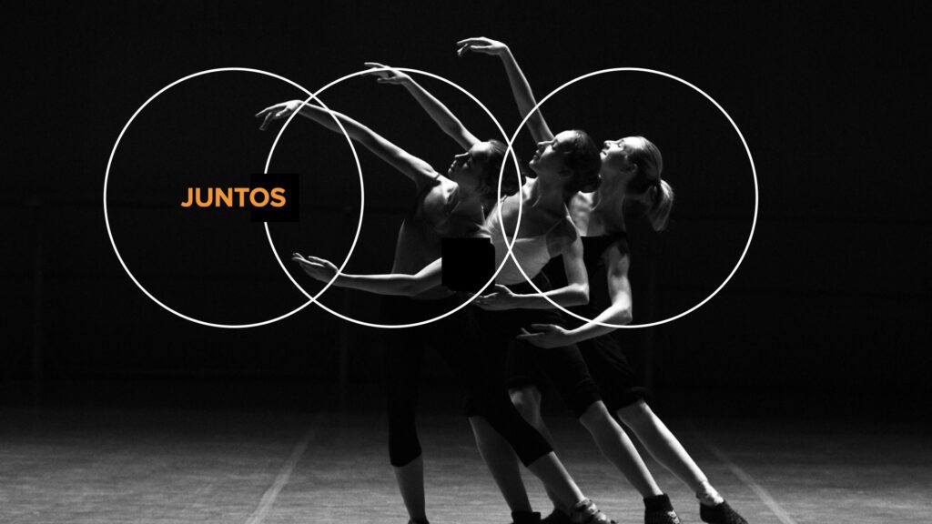 Fotografia artística em preto e branco de três bailarinas em cena, capturadas em um movimento coordenado e fluido. Elas estão levemente inclinadas para trás com os braços estendidos em poses elegantes, criando uma sobreposição visual de silhuetas sob uma iluminação dramática de palco. À esquerda, a palavra "JUNTOS" destaca-se em uma cor laranja vibrante, contrastando com o ambiente escuro e minimalista do estúdio de dança.