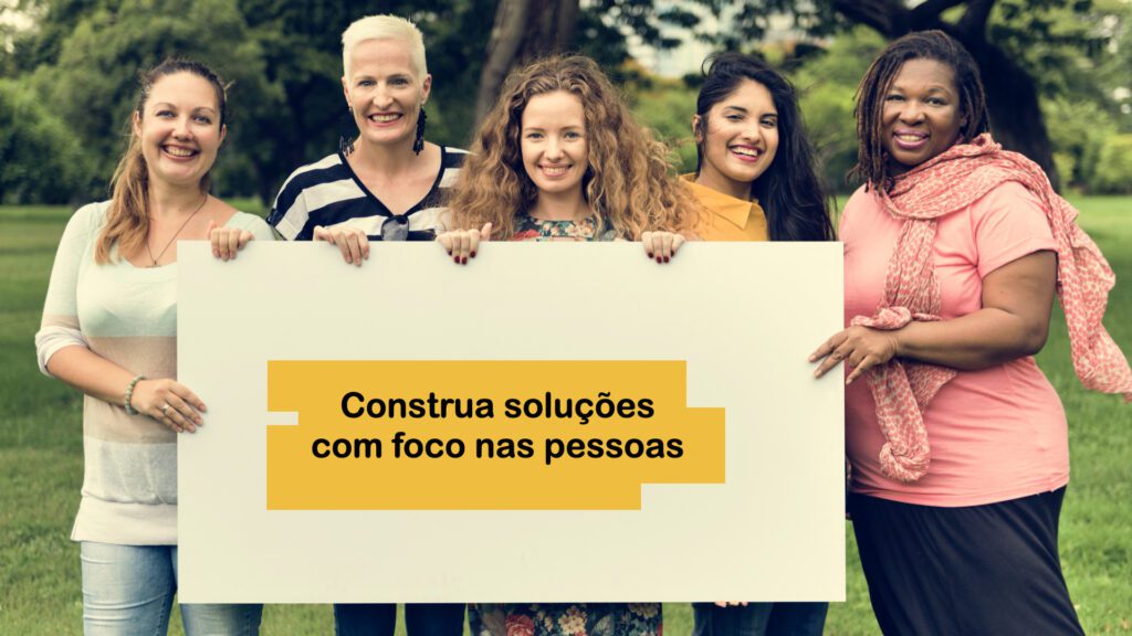 Cinco mulheres sorridentes em um parque segurando um grande cartaz branco. No centro do cartaz, há um retângulo amarelo com o texto em preto: "Construa soluções com foco nas pessoas".