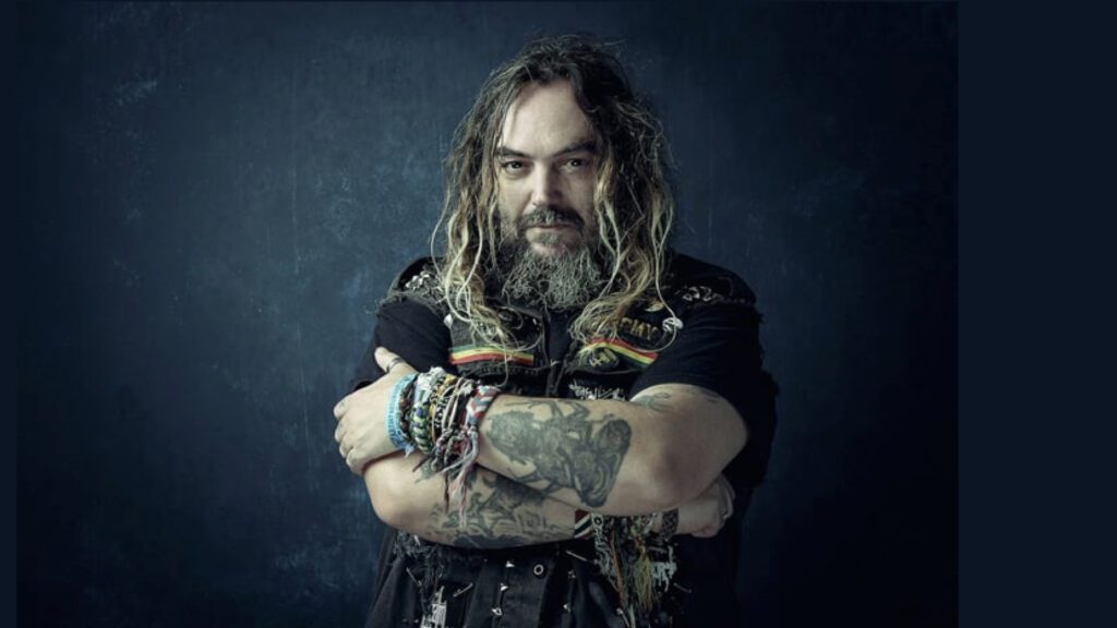 Retrato frontal do músico Max Cavalera em um fundo azul texturizado. Ele aparece de braços cruzados, exibindo tatuagens e diversos braceletes coloridos. Com cabelos longos, barba grisalha e vestindo um colete preto com patches, sua imagem simboliza o paralelo entre inovação e raízes culturais apresentado no World Creative Day 2023 em Santarém, Pará.