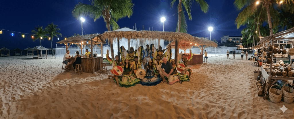 Fotografia noturna de um bar temático em Santarém, Pará, durante o World Creative Day 2023. No centro, dançarinos com roupas típicas verdes e amarelas e adereços de cabeça posam no chão de areia do local. Um homem caucasiano está agachado junto ao grupo. Ao redor, veem-se estruturas de madeira com teto de palha, coqueiros iluminados e uma exposição de cestaria e artesanato regional à direita.